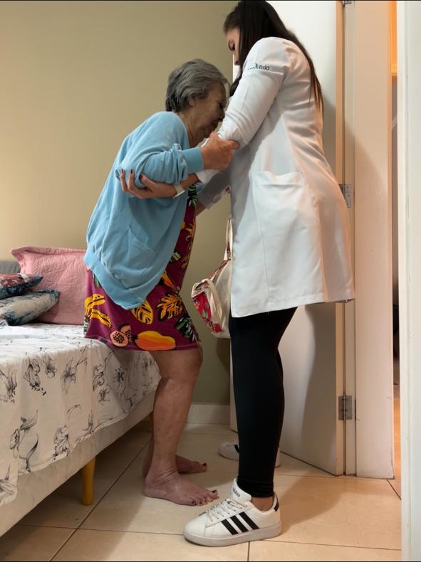 Bruna Kramer auxiliando paciente idosa a se levantar durante atendimento domiciliar em Valinhos