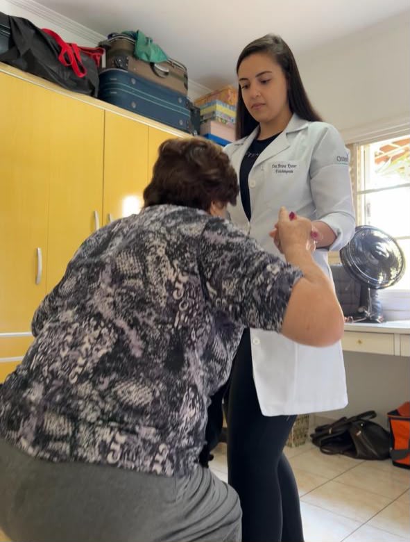 Bruna Kramer auxiliando paciente idosa em atendimento de fisioterapia geriátrica domiciliar